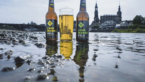 Zwei Lohrmanns-Bierflaschen und ein gefülltes Glas stehen im seichten Elbwasser vor der Stadtsilhouette Dresdens.