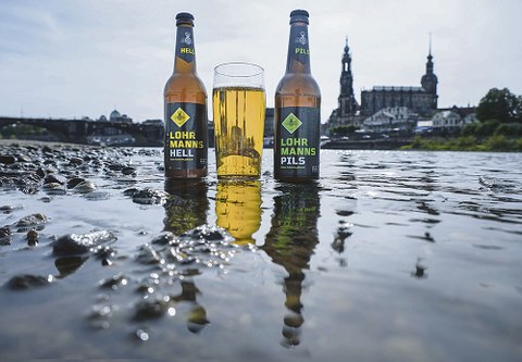 Zwei Lohrmanns-Bierflaschen und ein gefülltes Glas stehen im seichten Elbwasser vor der Stadtsilhouette Dresdens.