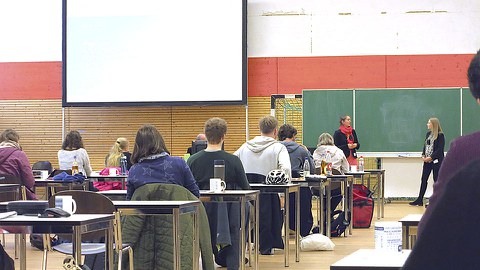 Teilnehmer an Tischen sitzend von hinten gesehen, vorne an Tafel zwei Frauen bei Präsentation.