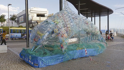 Überdimensionaler Delfin aus Plastikflaschen steht auf einer Uferpromenade.