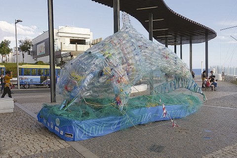 Überdimensionaler Delfin aus Plastikflaschen steht auf einer Uferpromenade.