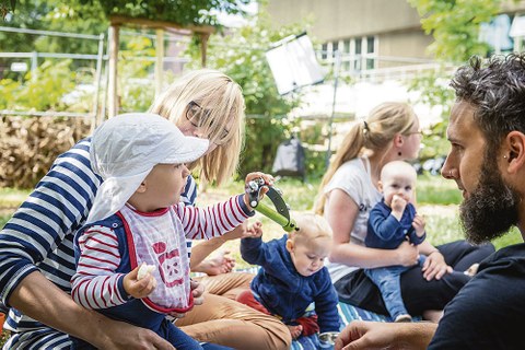 Kindergartenkinder sitzen mit Erwachsenen auf einer Wiese im Sommer.