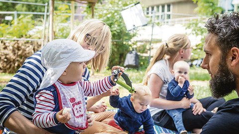 Kindergartenkinder sitzen mit Erwachsenen auf einer Wiese im Sommer.