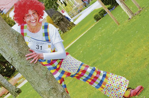 Der bunt gekleidete Clown Zitzewitz schaut hinter einem Baumstamm hervor.