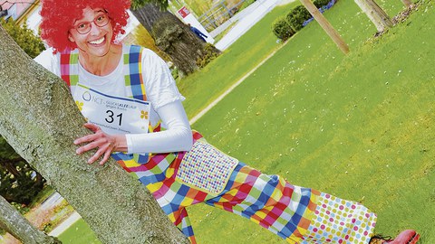 Der bunt gekleidete Clown Zitzewitz schaut hinter einem Baumstamm hervor.