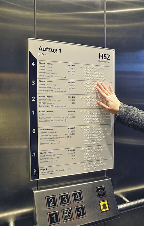 Zu sehen ist eine Tafel im Aufzug, auf der neben den Bezeichnungen der Stockwerke in Buchstaben auch die Bezeichnung in Braille-Schrift aufgebracht ist.