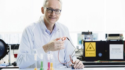 Prof. Leo sitzt an einem Labortisch. Im Hintergrund sind verschiedene Geräte zu sehen. Er hält eine Leuchtdiode in der Hand.