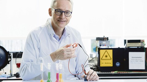 Prof. Leo sitzt an einem Labortisch. Im Hintergrund sind verschiedene Geräte zu sehen. Er hält eine Leuchtdiode in der Hand.