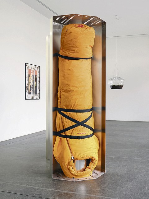 Ein gelber Schlafsack ist in einem offenen Metallkubus befestigt. Er hängt kopfüber.