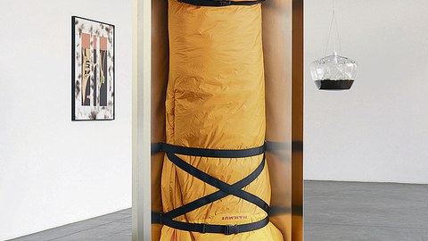 Ein gelber Schlafsack ist in einem offenen Metallkubus befestigt. Er hängt kopfüber.