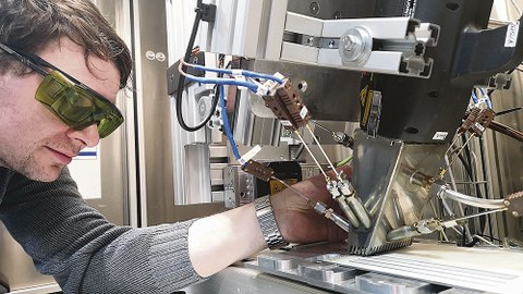 Techniker Erik Findeisen positioniert Thermoelemente an einem Laserkopf im Laserlabor des Instituts für Energietechnik an der TU Dresden. Darunter ist eine lackierte Probe zu sehen, die den Schutzanstrichen in alten Kernkraftwerken ähnelt.