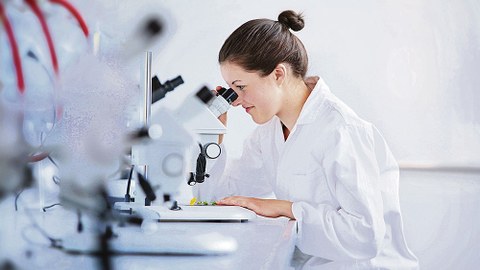 Eine Wissenschaftlerin in weißem Kittel sitzt an einem Tisch. Sie schaut in ein Mikroskop.