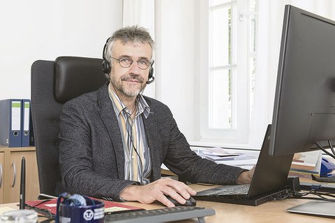 Prof. Kobel sitzt am Schreibtisch und schaut in die Kamera. Vor ihm steht ein aufgeklapptes Laptop, er hat Kopfhörer auf.