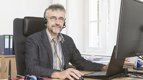 Prof. Kobel sitzt am Schreibtisch und schaut in die Kamera. Vor ihm steht ein aufgeklapptes Laptop, er hat Kopfhörer auf.