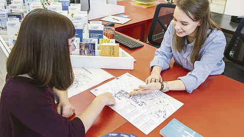 An einem Schreibtisch sitzen sich zwei Frauen gegenüber. Die eine erklärt der anderen den Campis-Plan der TUD.