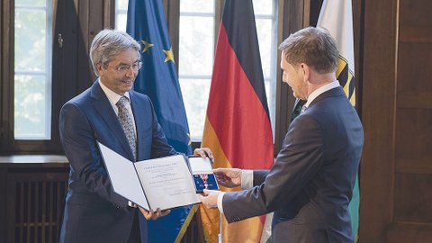 Sachsens Ministerpräsident Michael Kretschmer (r.) überreicht das Bundesverdienstkreuz und die Urkunde an Prof. Hans Müller-Steinhagen. Beide stehen vor den Flaggen Deutschlands, der EU und Sachsens.