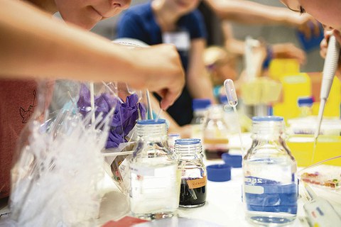 Glas-Laborbehältnisse stehen auf einem Tisch. Daraus werden von verschiedenen Menschen mit Pipetten Flüssigkeiten entnommen.