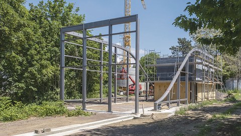 Die Stahl-Glas-Konstruktion steht auf dem Baugrundstück. Im Hintergrund rechts steht die zweisöckige BOX.