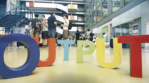 Die Buchstaben OUTPUT stehen auf dem Boden im Gebäude der Informatik-Fakultät. Im Hintergrund stehen Menschen im Gespräch.