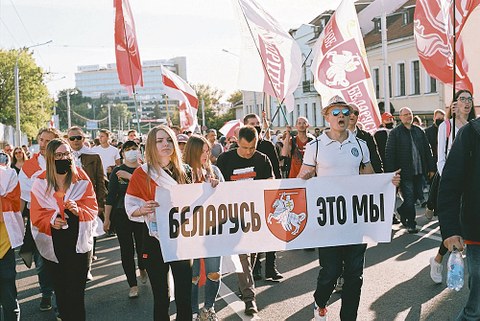 Menschen mit rot-weißen Fahnen laufen in einem Demonstrationszug auf der Straße. Im Vordergrund tragen eine junge Frau und ein Mann ein Transparent, auf dem in kyrillischen Buchstaben "Belarus, das sind wir" zu lesen ist.