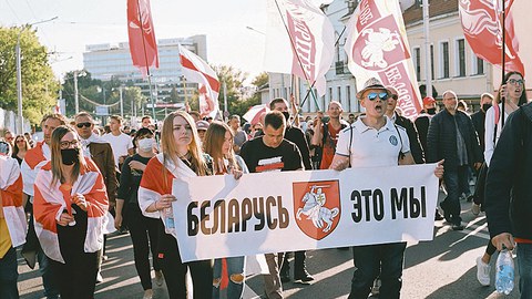 Menschen mit rot-weißen Fahnen laufen in einem Demonstrationszug auf der Straße. Im Vordergrund tragen eine junge Frau und ein Mann ein Transparent, auf dem in kyrillischen Buchstaben "Belarus, das sind wir" zu lesen ist.