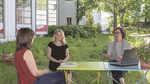 Die drei Frauen sitzen sich zugewandt an einem Gartentisch, auf dem ein Buch und ein Laptop stehen, im Gemeinschaftsgarten auf der Wiese.