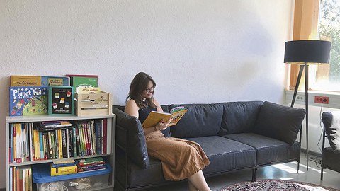 Tina Czaja sitzt auf einem schwarzen Sofa. Sie schaut in ein aufgeschlagenes Buch, das sie in der Hand hält. Links neben dem Sofa steht ein Regal voller Bücher. Rechts neben dem Sofa steht eine Lampe. Vor dem Sofa liegt ein runder Teppich.