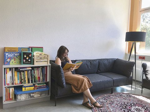 Tina Czaja sitzt auf einem schwarzen Sofa. Sie schaut in ein aufgeschlagenes Buch, das sie in der Hand hält. Links neben dem Sofa steht ein Regal voller Bücher. Rechts neben dem Sofa steht eine Lampe. Vor dem Sofa liegt ein runder Teppich.