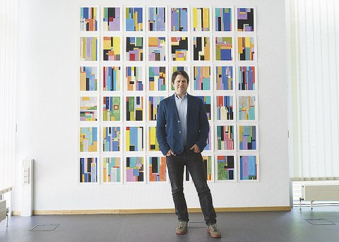 Anton Ginzburg steht vor seiner Serie. Diese besteht aus acht mal sechs kleinformatigen Bildern. Die Bilder sind geometrisch und bunt.