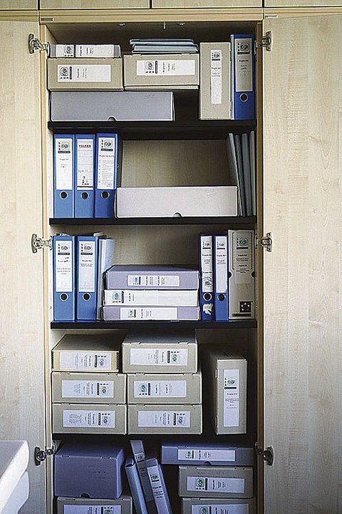 Zu sehen ist ein geöffneter Aktenschrank, in dem blaue Aktenordner und graufarbene Kisten stehen oder liegen.