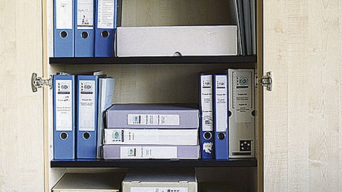 Zu sehen ist ein geöffneter Aktenschrank, in dem blaue Aktenordner und graufarbene Kisten stehen oder liegen.