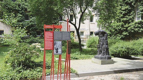 Links neben der Wieland-Förster-Plastik stehen auf roten Streben rote und graue Tafeln mit Texten.