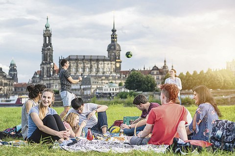 Eine Gruppe junger Menschen sitzt auf einer Wiese am Elbufer. Im Hintergrund spielen ein Mann und eine Frau Ball. Zu sehen ist die Silhouette Dresdens mit Hofkirche, Schloss und Italienischem Dörfchen.