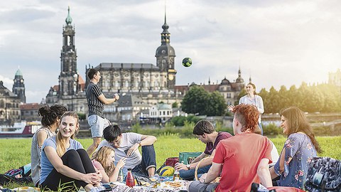 Eine Gruppe junger Menschen sitzt auf einer Wiese am Elbufer. Im Hintergrund spielen ein Mann und eine Frau Ball. Zu sehen ist die Silhouette Dresdens mit Hofkirche, Schloss und Italienischem Dörfchen.