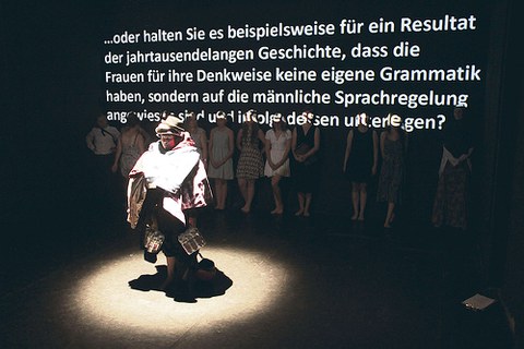 Eine Person steht im Scheinwerferlicht vor einer Reihe Menschen. Darüber ist ein Text zu lesen: ... oder halten Sie es beispielsweise für ein Resultat der jahrtausendelangen Geschichte, dass die Frauen für ihre Denkweise keine eigene Grammatik haben, ...