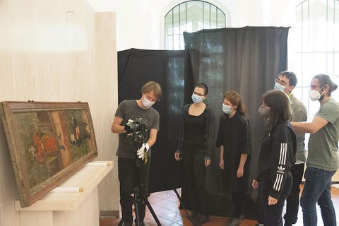 Ein Bildnis ist auf einem Tisch aufgestellt und an die Wand gelehnt. Davor stehen fünf Personen. Eine Person betreut einen Fotoapparat, der auf einem Stativ vor dem Tisch steht.