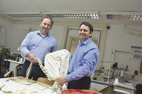 Dr. Andreas Haka und Prof. Yordan Kyosev stehen in einem Laborraum. Sie halten einen hellen Fallschirmstoff in den Händen.