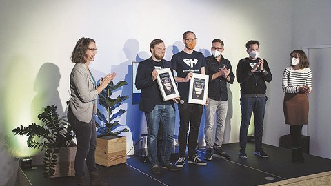Die Gewinner des Battles Ilja Bauer und sein Team von EntwicklerHeld stehen auf einer Bühne und halten die Urkunden in der Hand.
