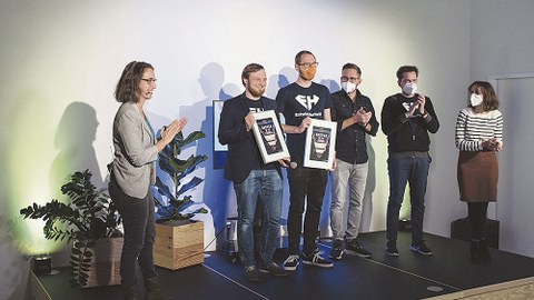 Die Gewinner des Battles Ilja Bauer und sein Team von EntwicklerHeld stehen auf einer Bühne und halten die Urkunden in der Hand.