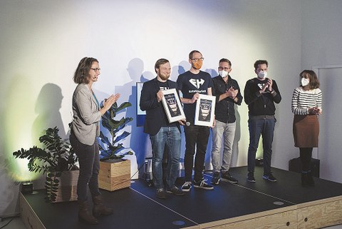 Die Gewinner des Battles Ilja Bauer und sein Team von EntwicklerHeld stehen auf einer Bühne und halten die Urkunden in der Hand.