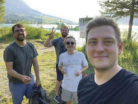 Assad Majid, Dennis Wenzel, Robert Bauer und Philipp Strietzel stehen am Ufer eines Sees und schauen lachend in die Kamera.