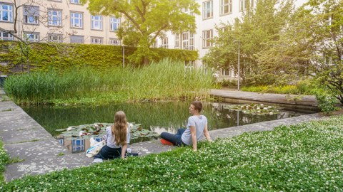Das Foto zeigt zwei Studierende an einem Teich hinter dem Barkhausen-Bau der TU Dresden. Sie sitzen nebeneinander auf dem Boden. Auf der großen Wiese im Vordergrund blüht weißer Klee.