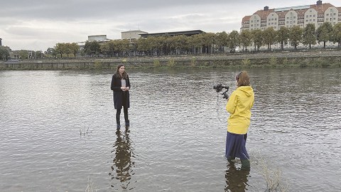 Beide Frauen stehen knöchelhoch im Wasser der Elbe in Dresden. Im Hintergrund ist am Ufer der Sächsische Landtag und ein Hotel zu sehen.