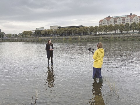 Beide Frauen stehen knöchelhoch im Wasser der Elbe in Dresden. Im Hintergrund ist am Ufer der Sächsische Landtag und ein Hotel zu sehen.