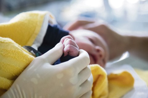 Zu sehen ist ein Neugeborenes, eingewickelt in eine gelbe Decke. Eine Hand mit weißem Handschuh hält die Hand des Babys.