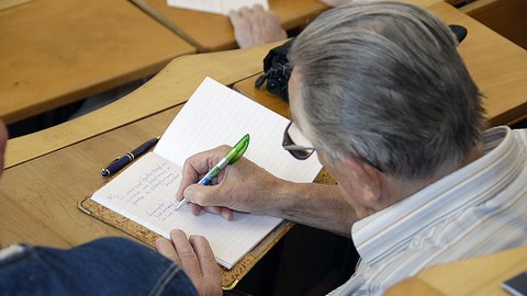 Auf dem Foto ist ein älterer Mann von hinten zu sehen, der in einem Hörsaal sitzt und in einem Heft schreibt.