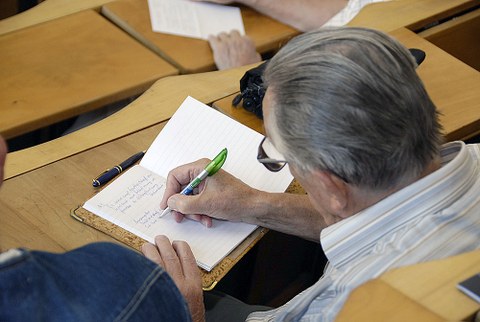 Auf dem Foto ist ein älterer Mann von hinten zu sehen, der in einem Hörsaal sitzt und in einem Heft schreibt.