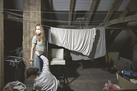 Auf einer Leine hängen Betttücher. Davor steht eine Frau mit Maske, ihr wird durch einen Mann das Bein vermessen.