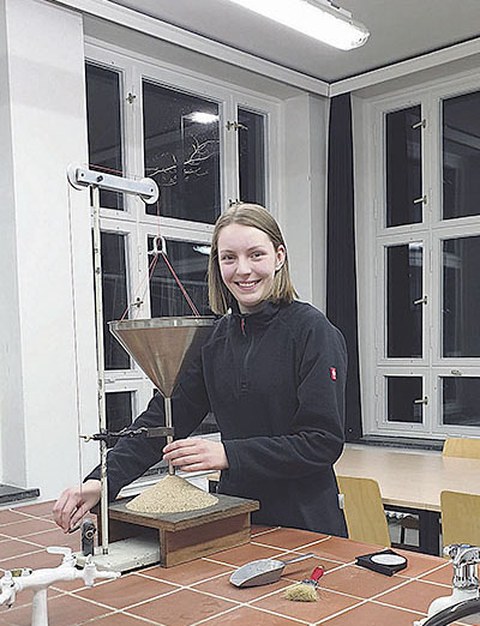 Sarah Müller steht hinter einem gekachelten Labortisch. Vor ihr ist ein Versuch aufgebaut. Aus einem Trichter läuft ein sandartiger Baustoff auf eine Unterlage.