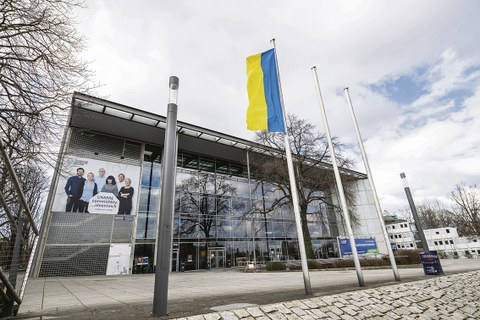 Das Foto zeigt das Hörsaalgebäude der TU Dresden. Davor stehen drei Fahnenmasten, an dem linken ist die Flagge der Ukraine gehisst.